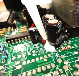 Hand-dispensing white sealant around electrolytic capacitors on PCB for enhanced mechanical bonding.